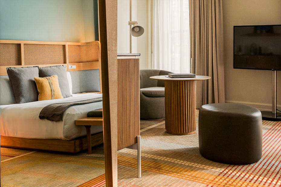 Junior Suite at Hotel Ändra with a modern bed, woven headboard, gray and mustard pillows, gray club chair, round wood table, leather ottoman, large window, and flat-screen TV.
