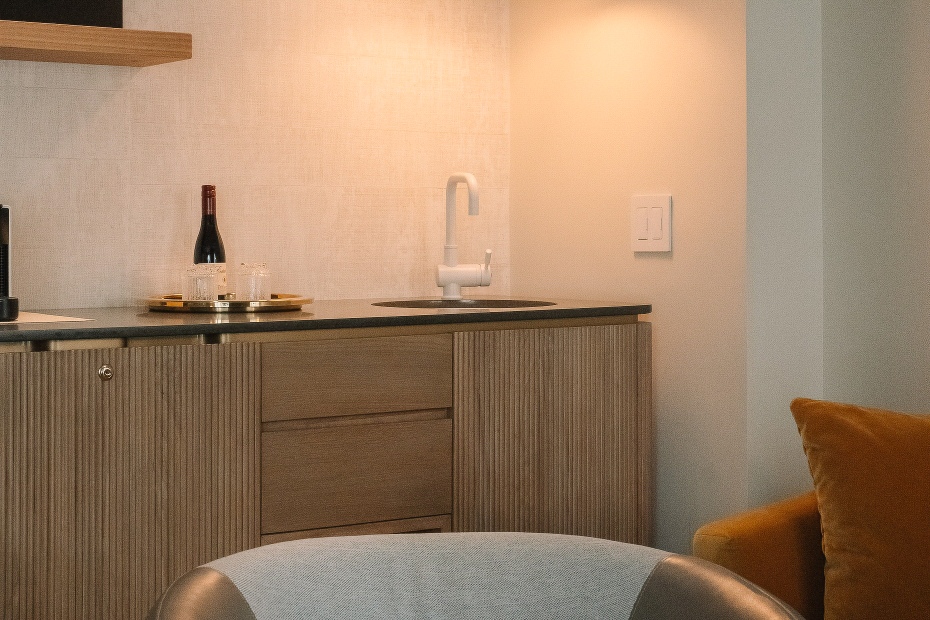 Junior Suite wet bar at Hotel Ändra with wood cabinets, white faucet and sink, a tray with a wine bottle and Scandinavian glasses, and part of a gray chair and mustard sofa